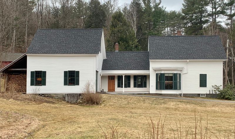 White farmhouse with dark shutters and roof, brown door, set in a field.