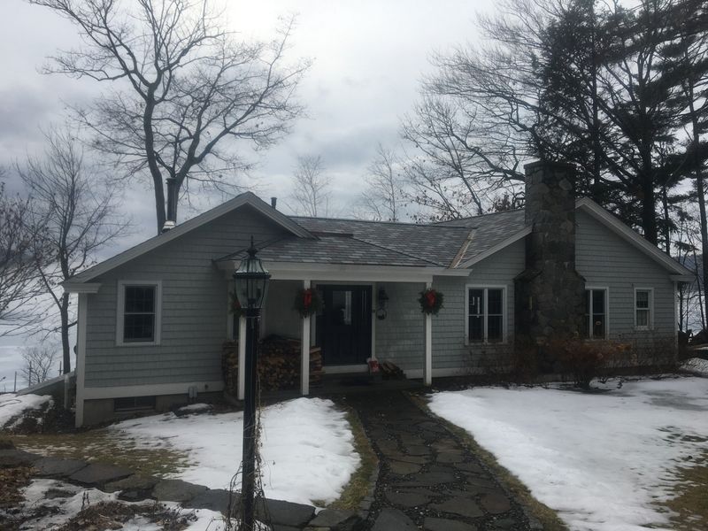A one-story light gray house with a stone chimney and walkway, surrounded by snow and bare trees.