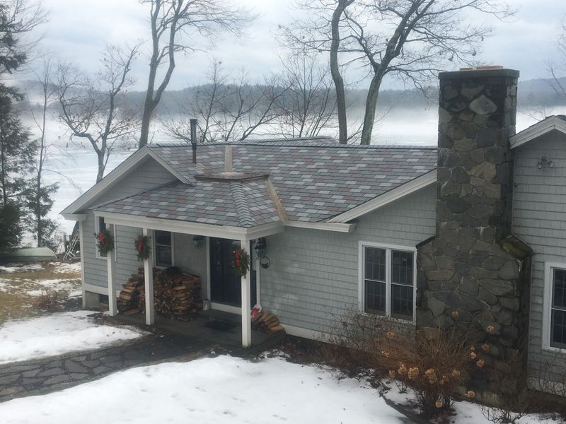 Grey house with stone chimney, snow, and foggy lake in the background.