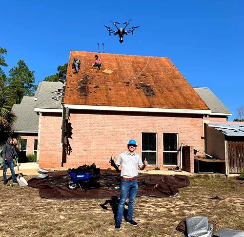 Workers repair a house roof, with a drone overhead and debris on the ground.