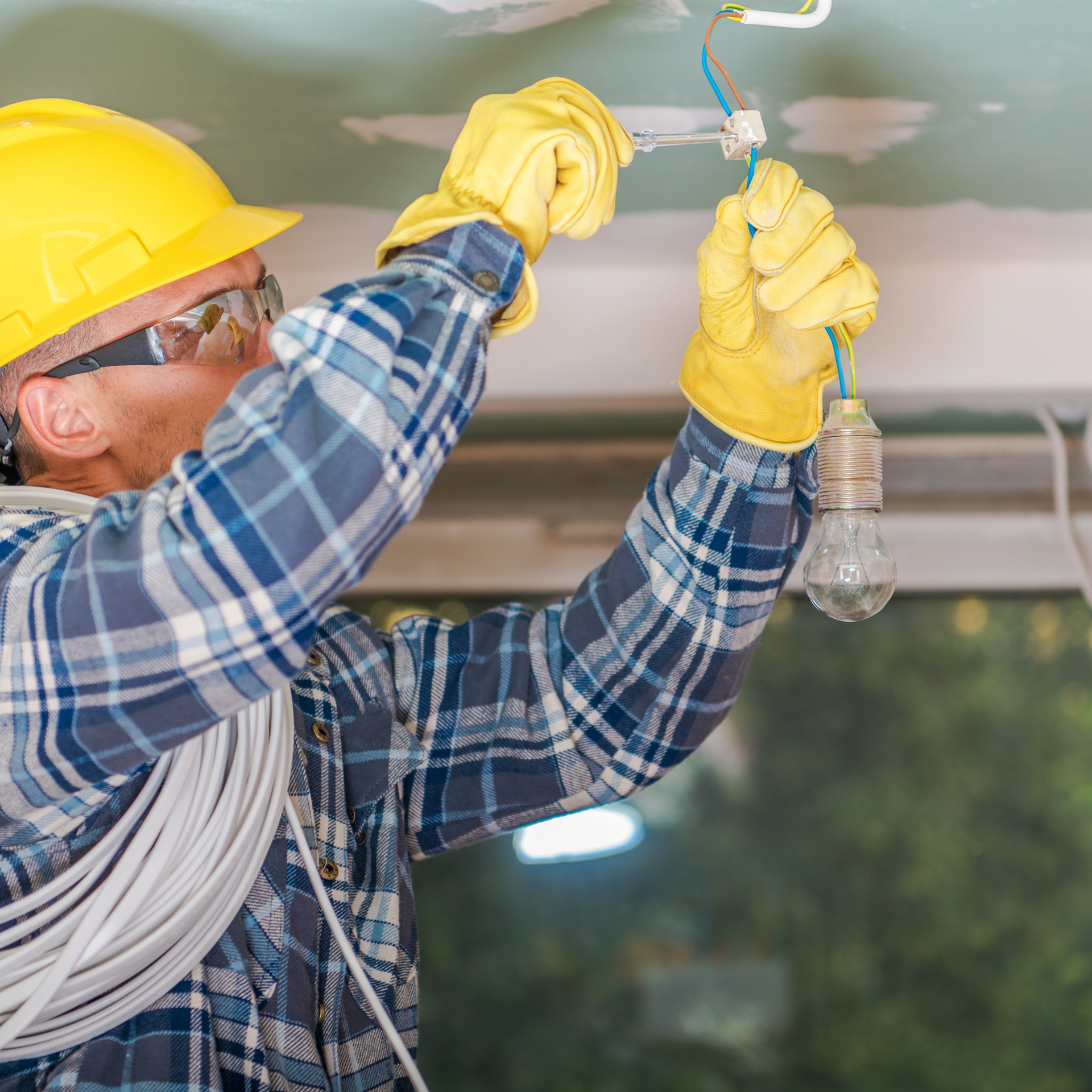 Electrician in a yellow hard hat and gloves connecting wires to a light bulb in a room.
