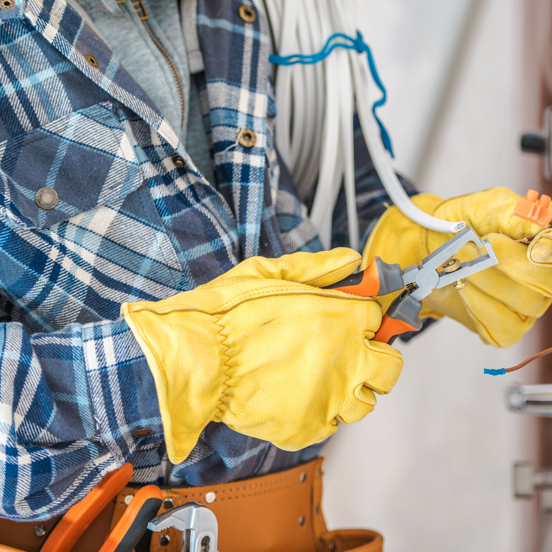 Electrician, gloved hands, stripping wire with pliers, plaid shirt, tool belt.