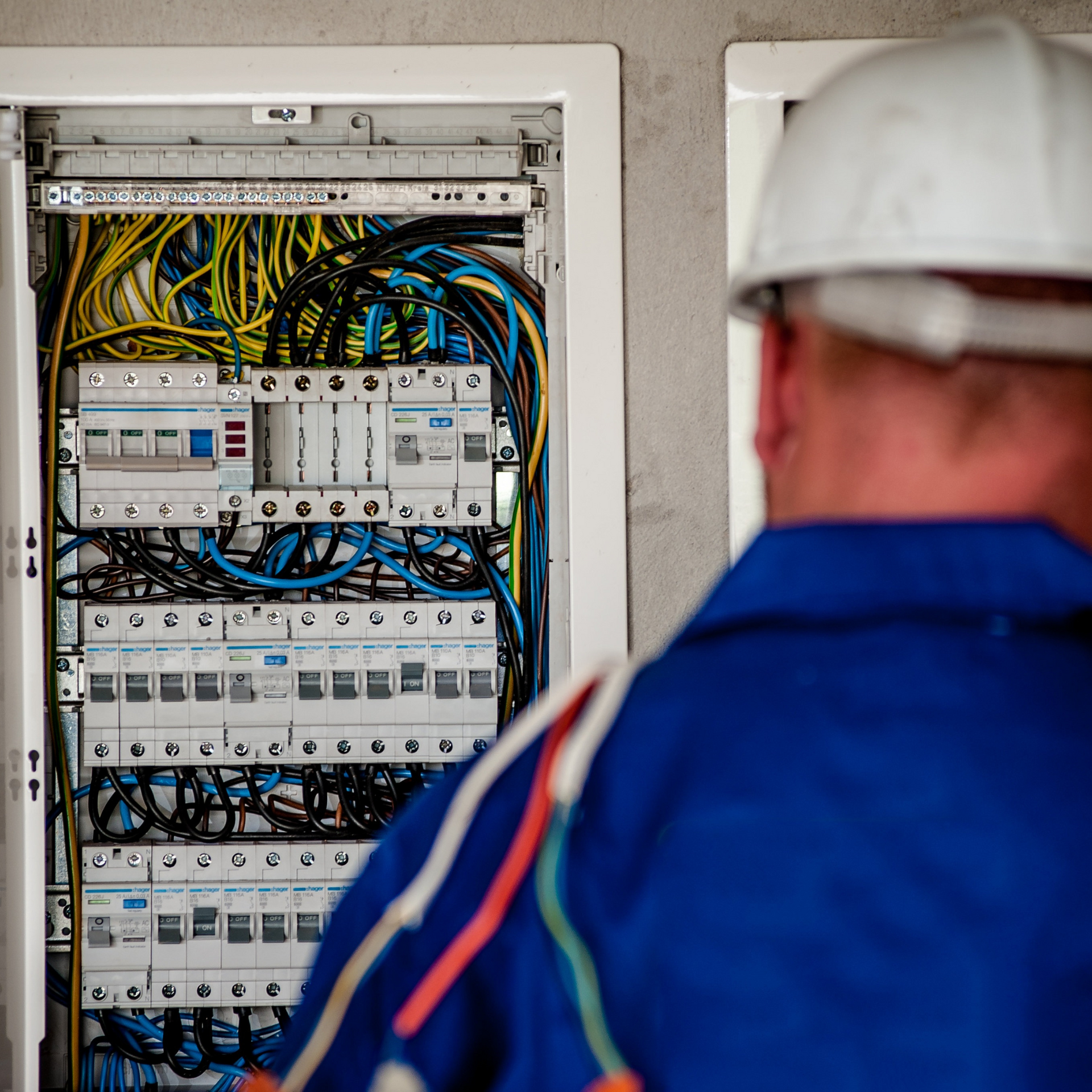 Electrician in blue uniform and hard hat inspecting an open electrical panel with colorful wires.