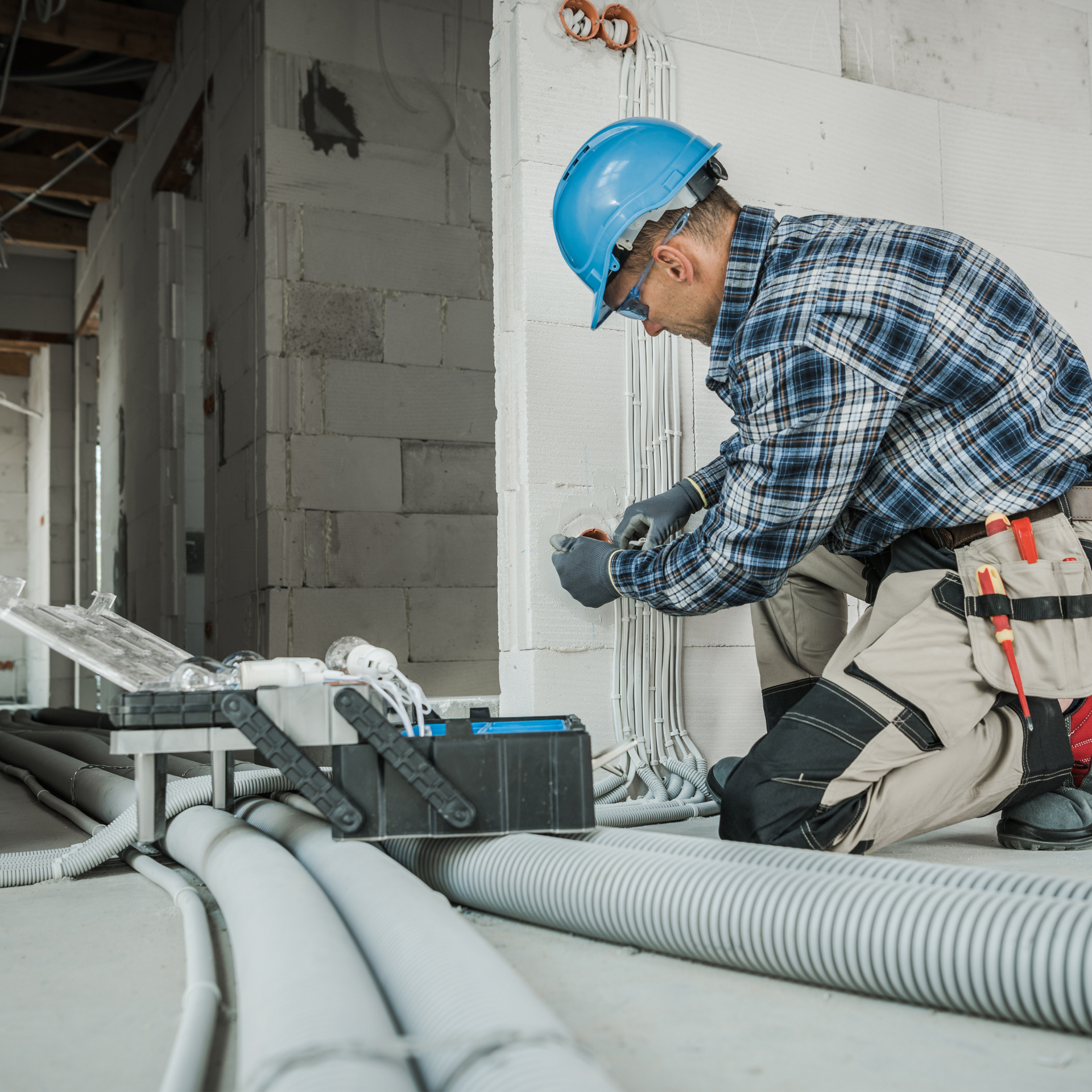 Electrician kneels, wiring a wall in a construction site; blue hard hat, plaid shirt, tools.