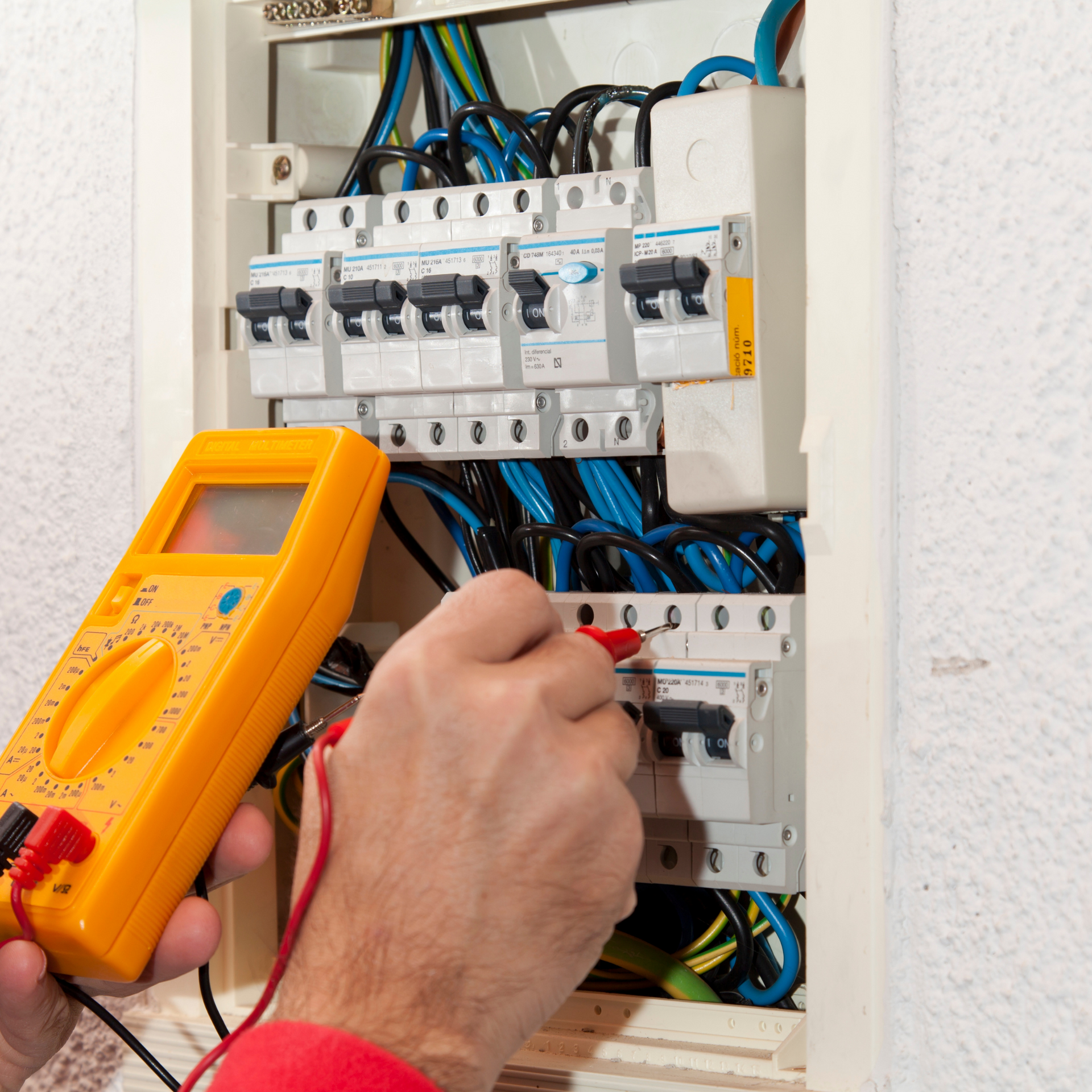 Electrician using a yellow multimeter to test electrical panel wiring in a white box.