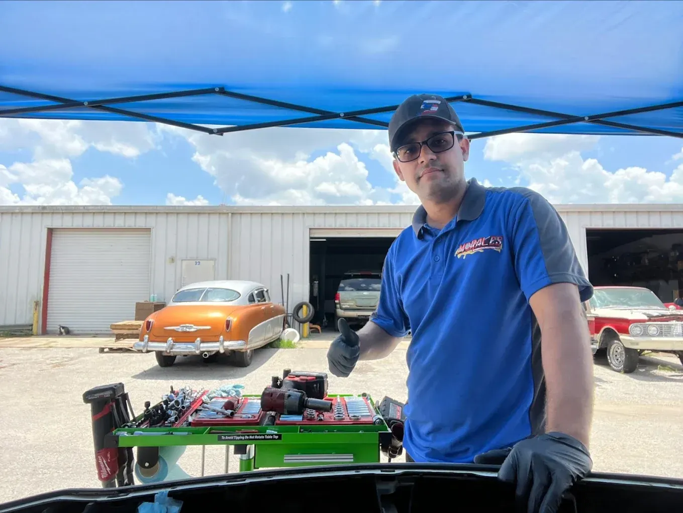 Man with thumbs-up in front of a classic car, tools, and a garage. Blue shirt, sunglasses, sunny outdoor setting.