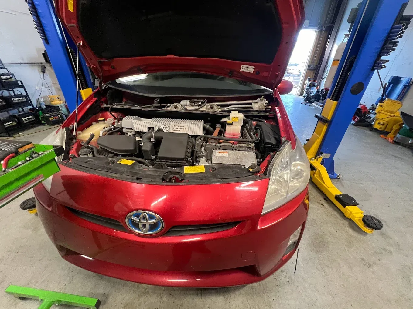 Red Toyota Prius with hood open in a repair shop, raised on a lift.