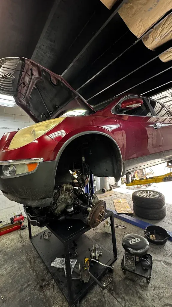 A maroon car being repaired in a garage, hood open. The front suspension is exposed on a lift.