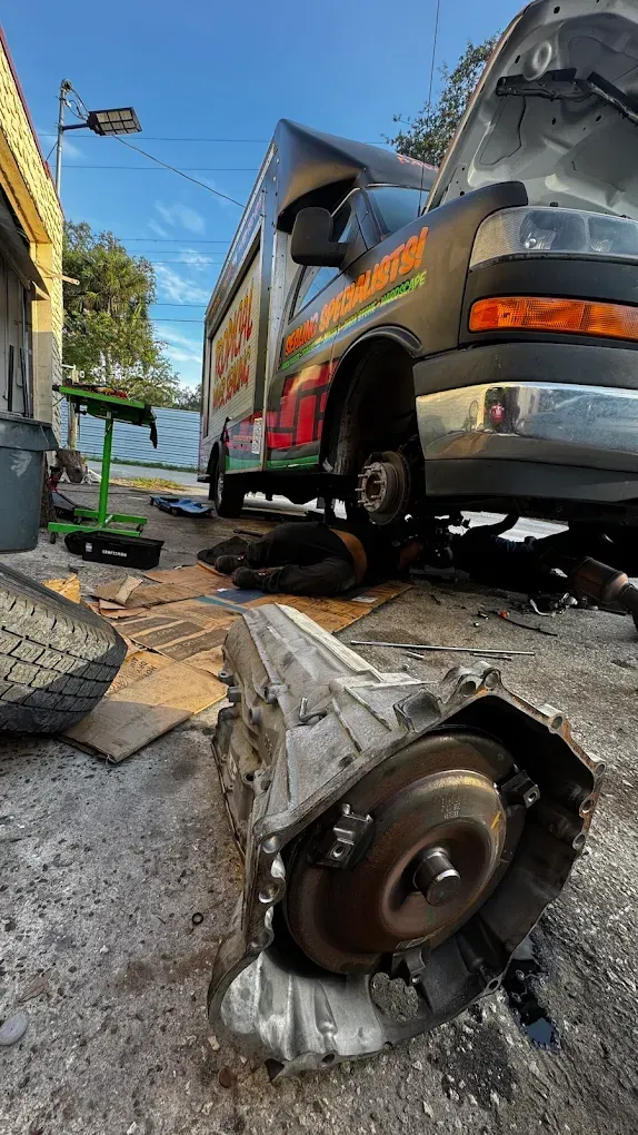 Mechanic working on a van, transmission on the ground. Outdoor repair shop setting.