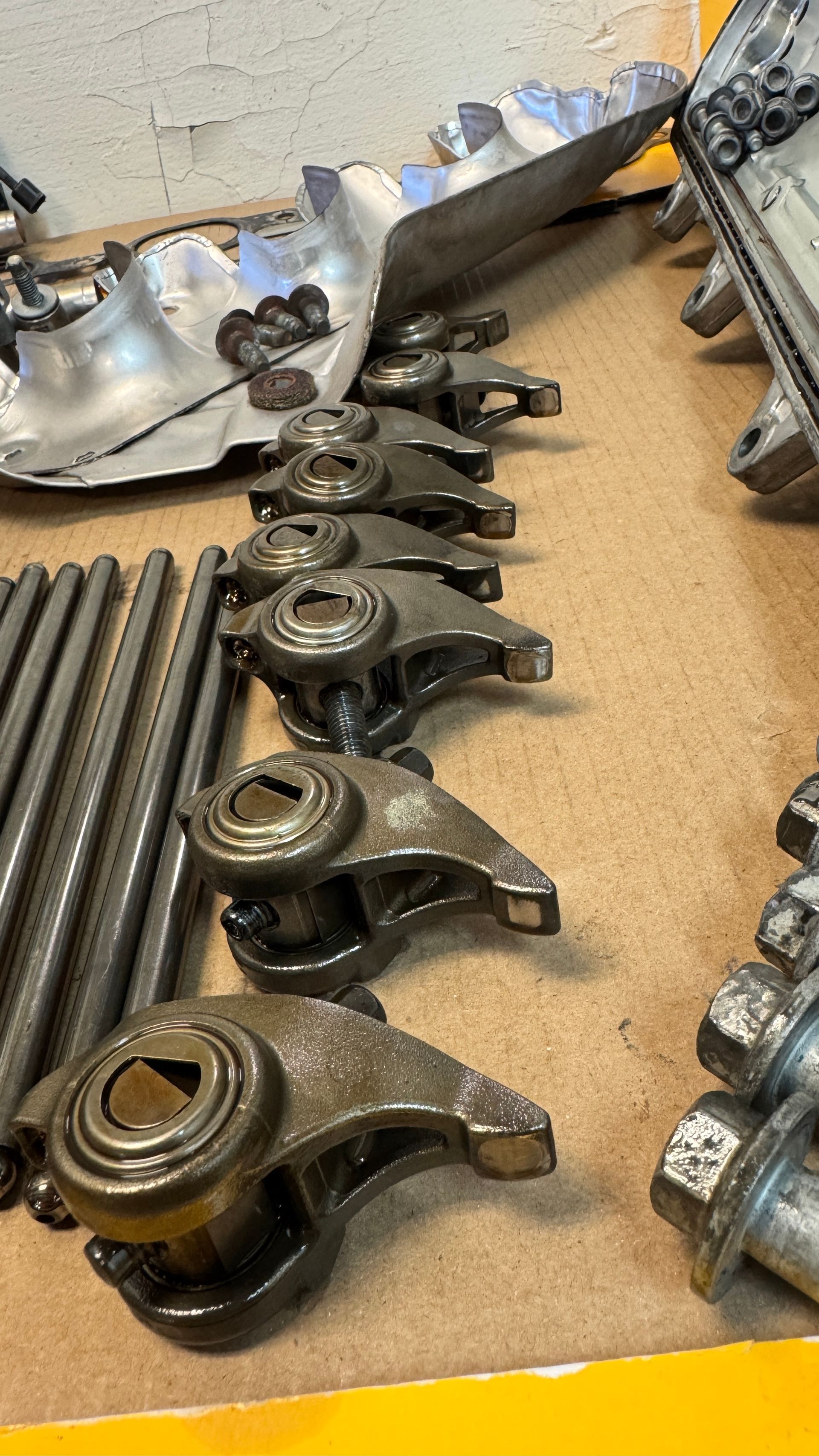Engine rocker arms and pushrods arranged on a workbench beside a cylinder head and various mechanical bolts.