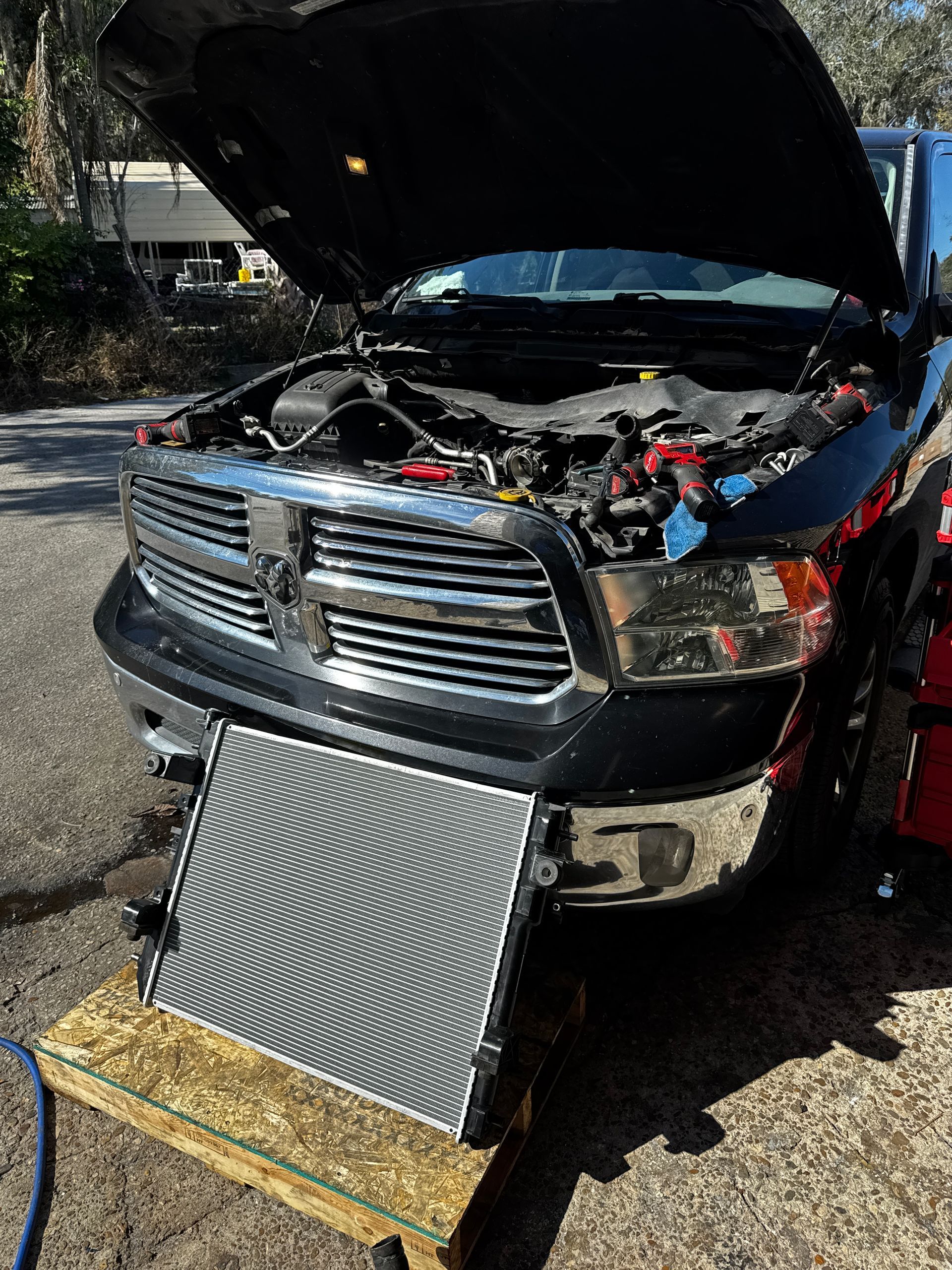 A black Ram pickup truck with an open hood and its new radiator sitting on a wooden stand in front of the front bumper.