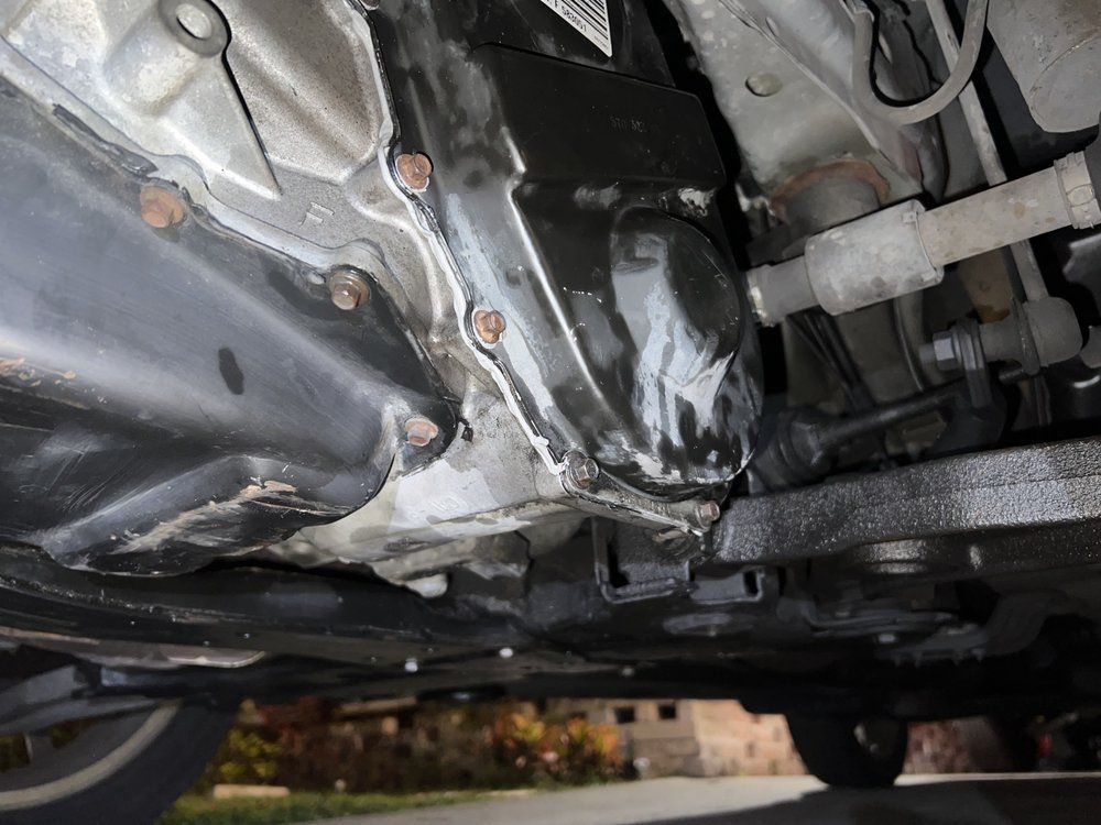 Underside of a car engine, showing an oil leak on the transmission pan and surrounding parts.