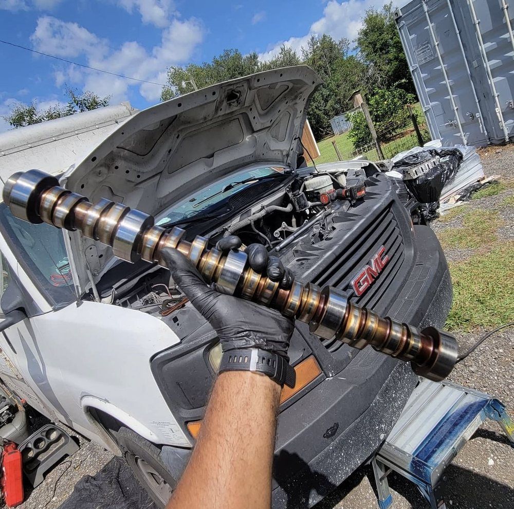 A hand wearing a black glove holding a camshaft in front of an open-hood GMC van.