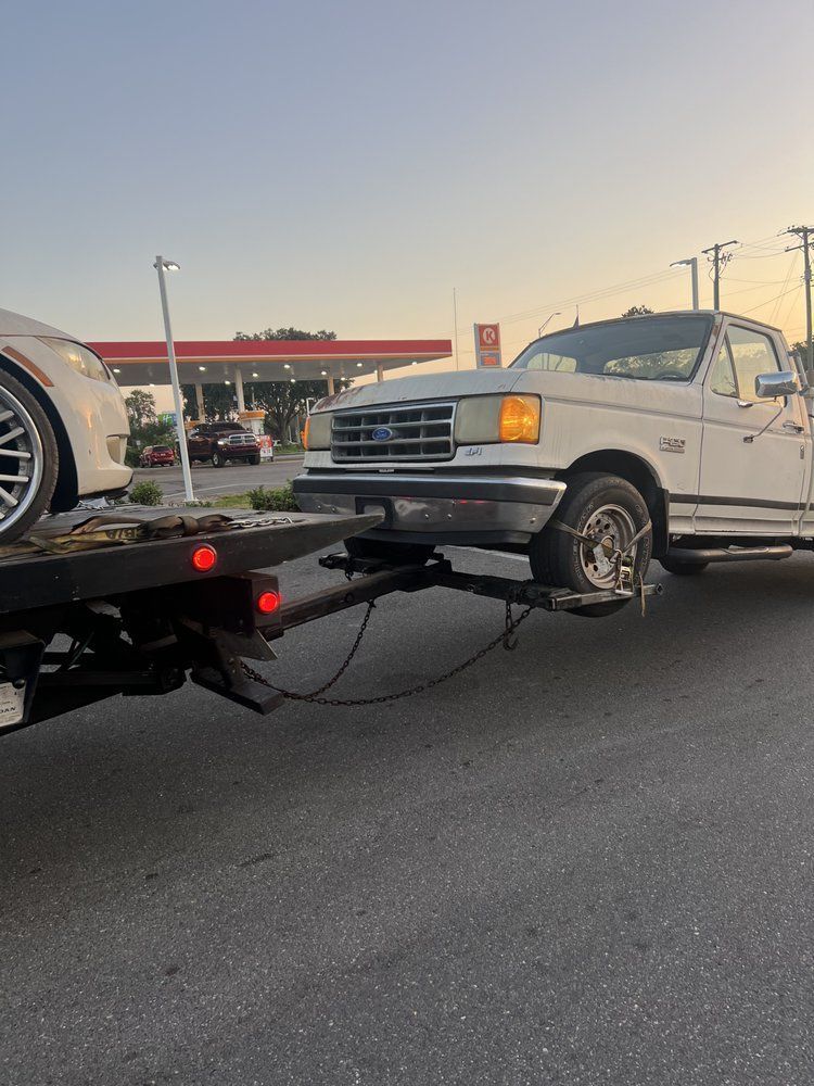 White tow truck hauling a car on a flatbed trailer, in front of a gas station at dusk.