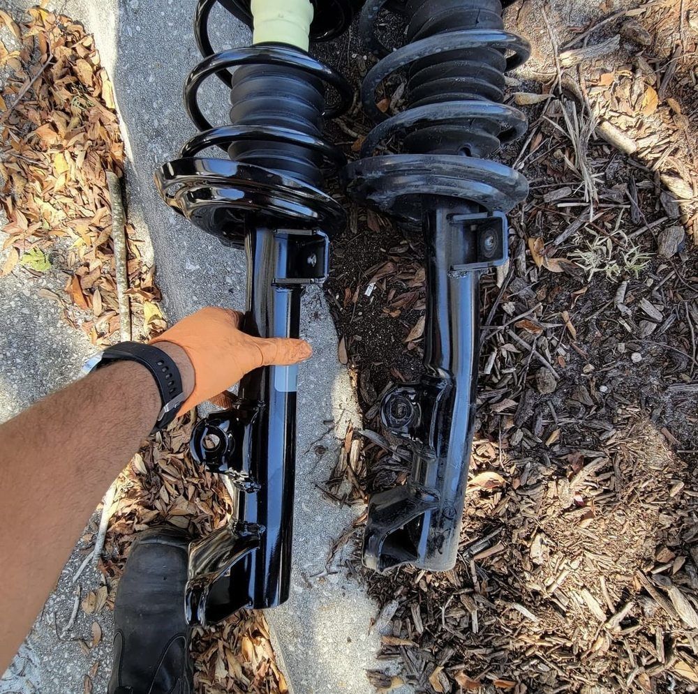Pair of black car struts on ground, one held by gloved hand.