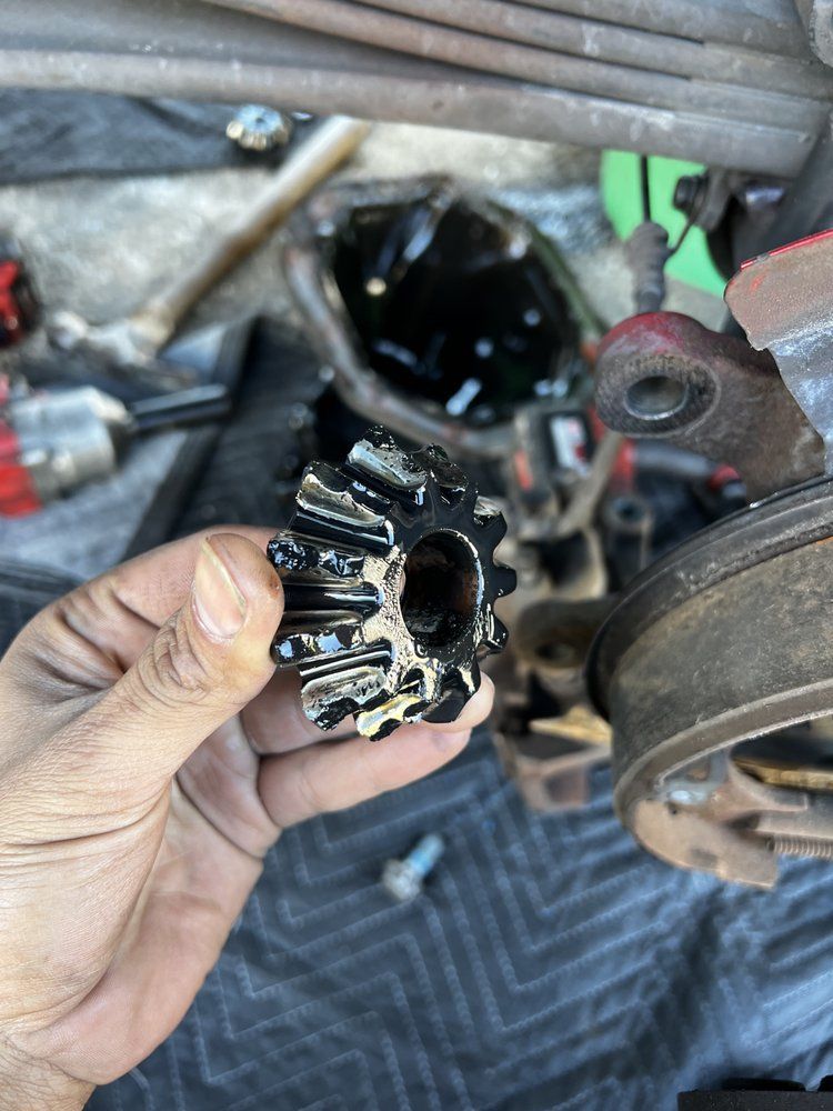 Hand holding a black gear removed from a vehicle's rear axle. Parts of a car are in the background.