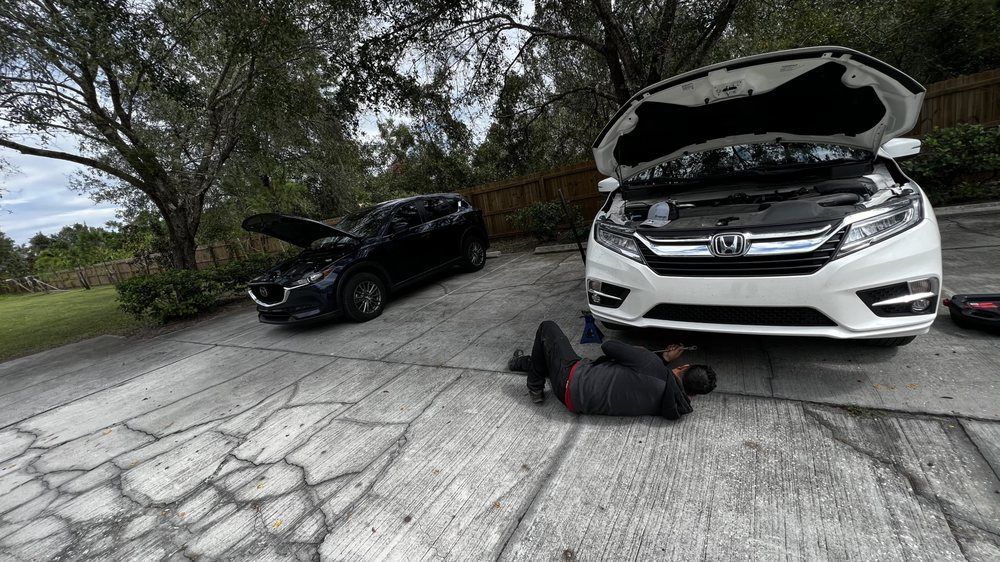 Person working on a white car with an open hood; black car in the background. Paved driveway, trees and sky.