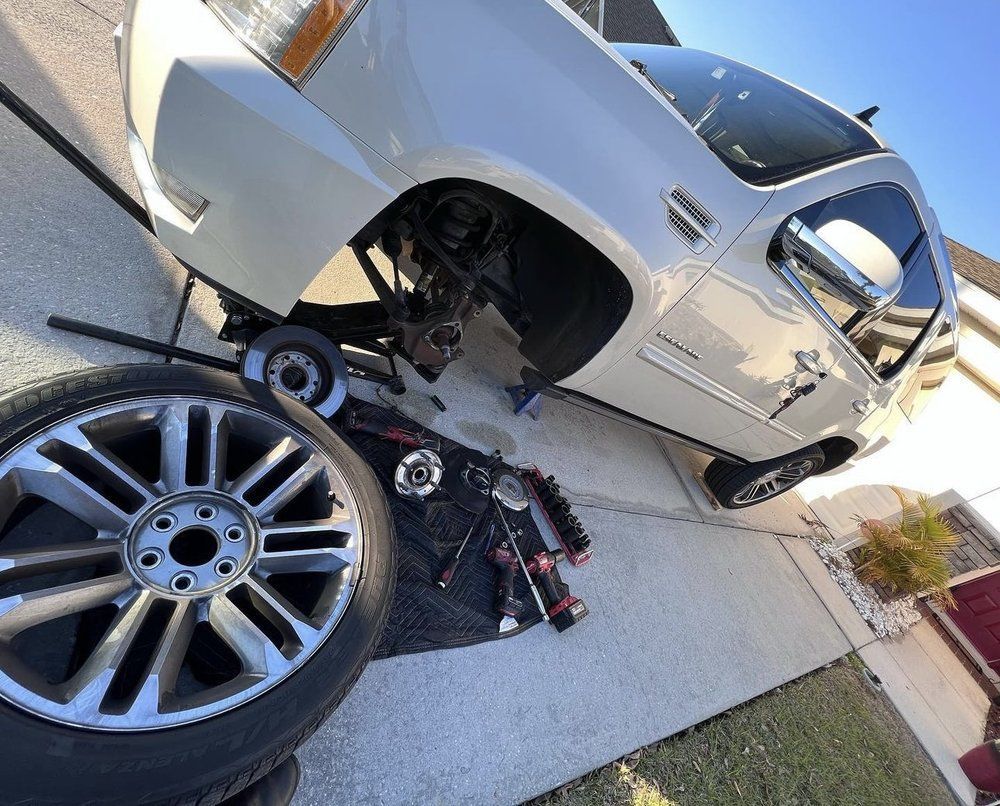 White Cadillac Escalade with front tire removed, surrounded by tools and spare tire in driveway.