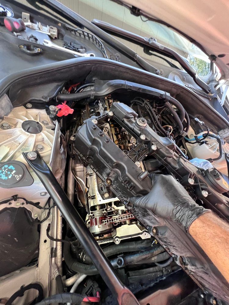 A person in gloves removing a black engine valve cover from a car. Open hood, engine bay visible.