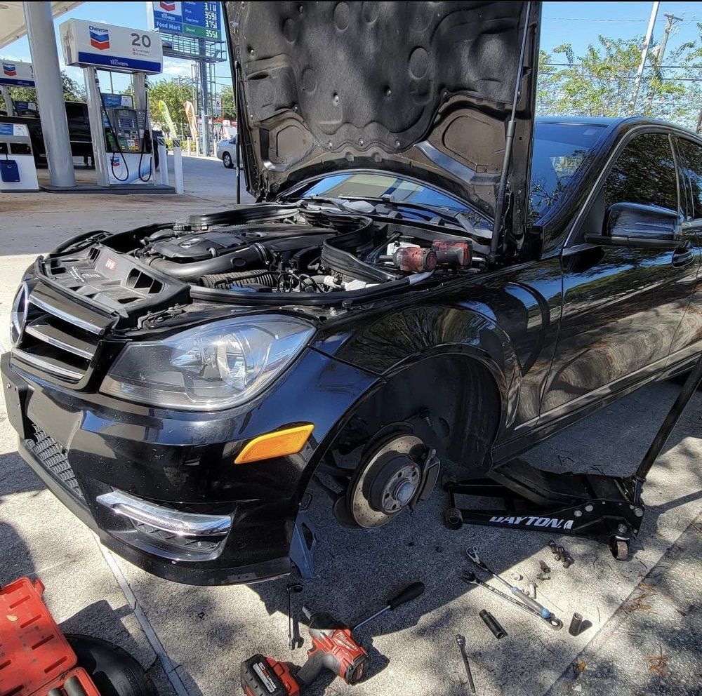 Black Mercedes with open hood and missing front tire at a gas station, with tools nearby.