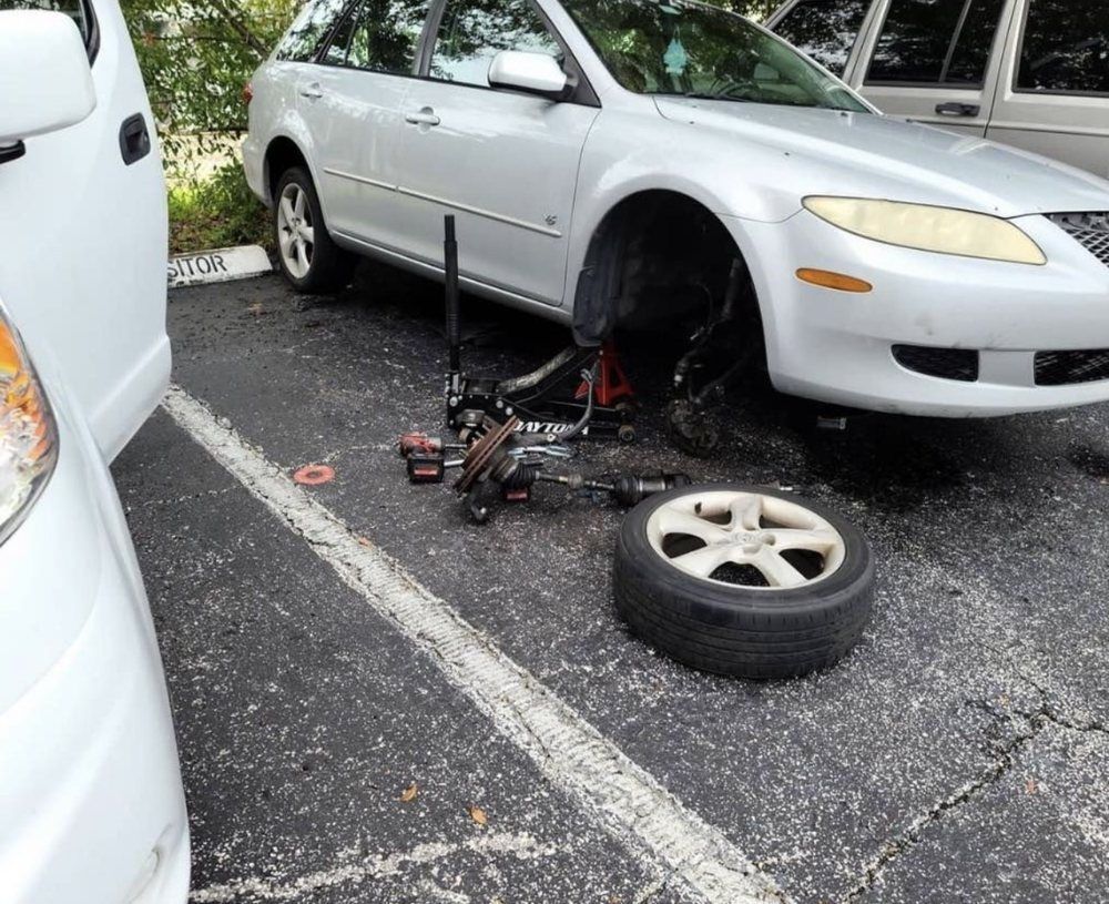 Car being worked on in parking lot; tire removed, jack in use. Silver vehicle next to white car.