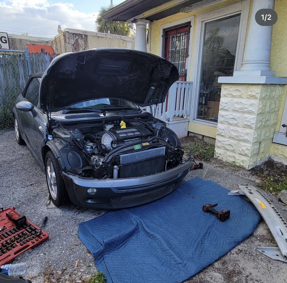 Black Mini Cooper with open hood, being worked on outside a yellow building.