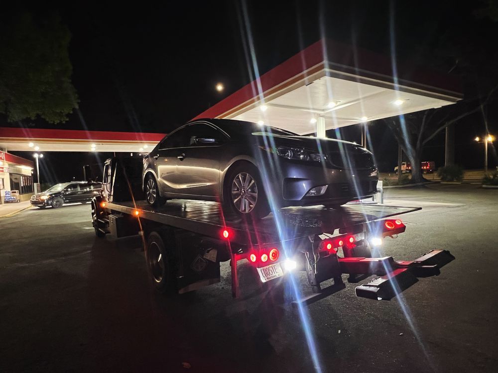 A tow truck carrying a silver car at night, under a gas station canopy.