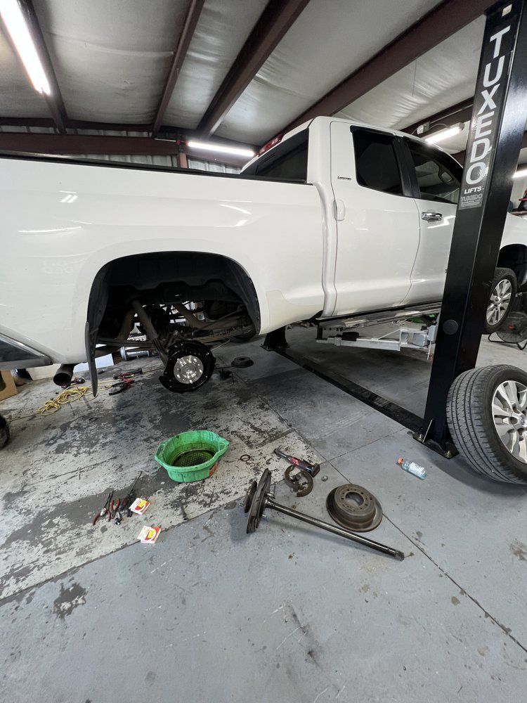 White truck on a lift with a removed wheel in a repair shop; tools and parts on the floor.