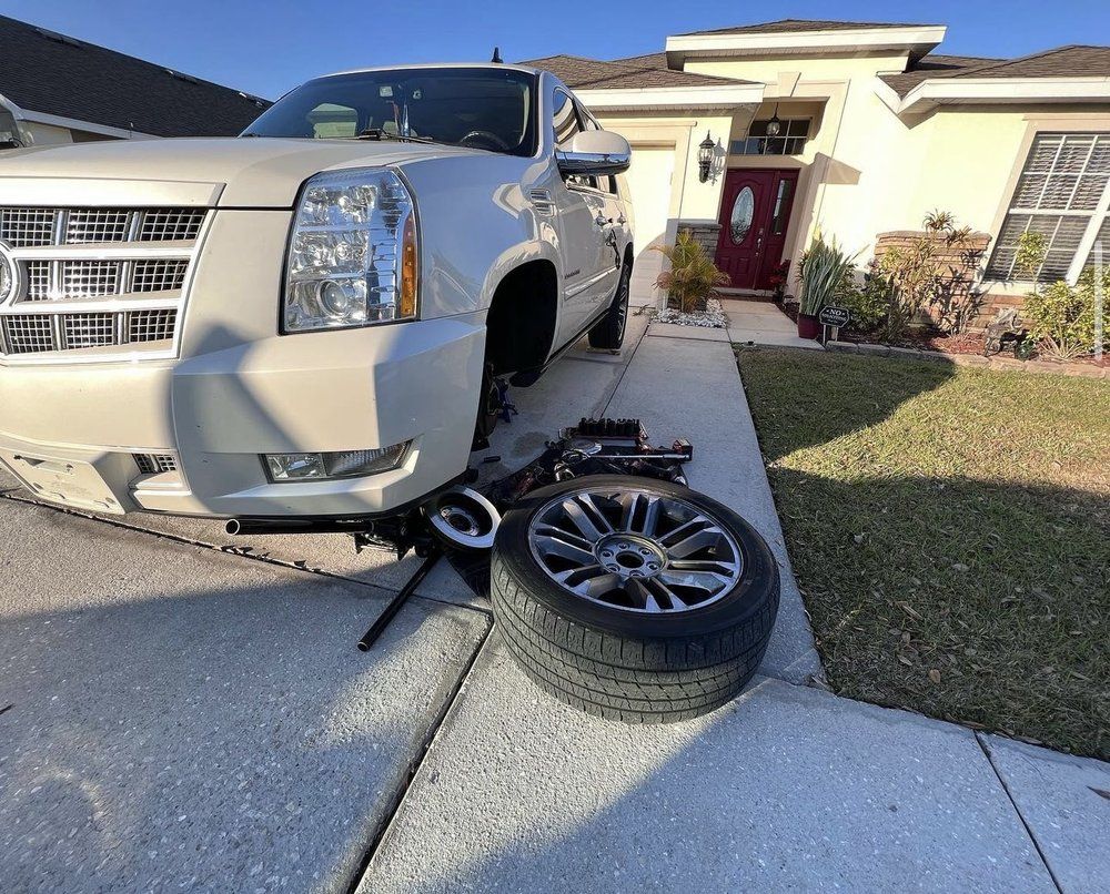 White SUV on a driveway with a tire removed, jack and tools present, in front of a house.