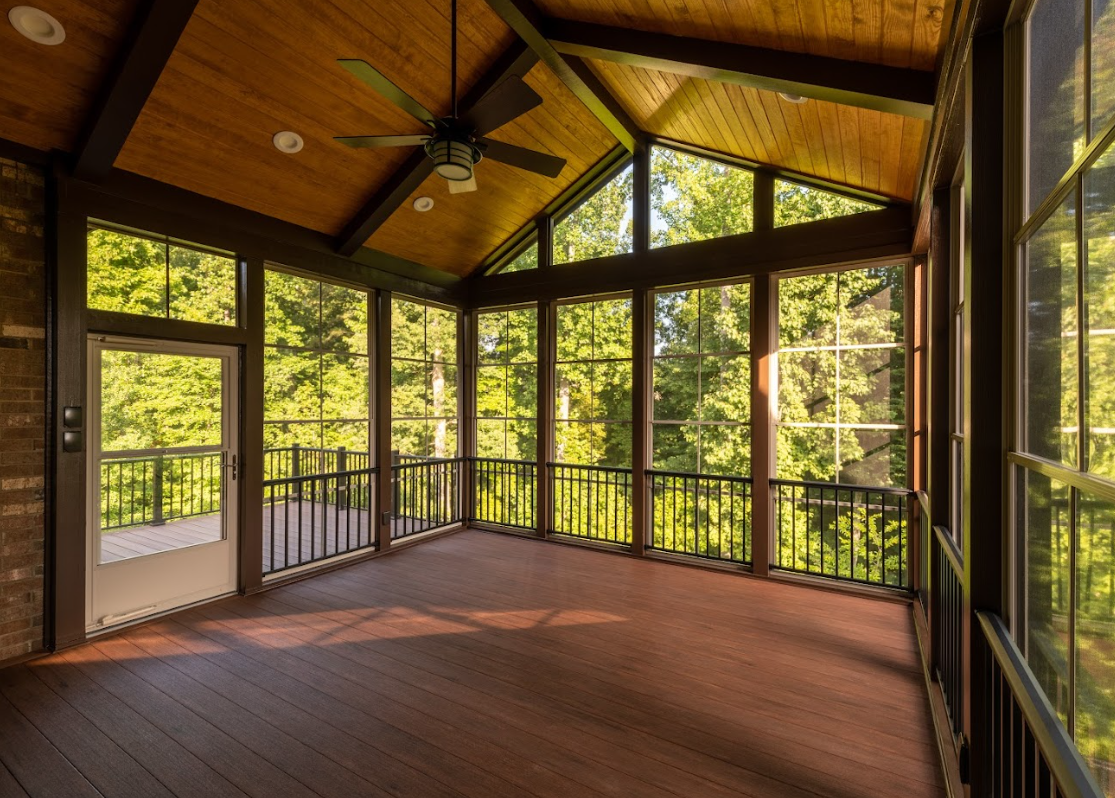 Screened-in porch with brown floor, wood ceiling, and tall windows overlooking a forest.