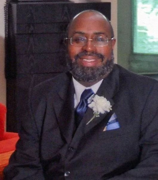 Man in a suit smiles, wearing glasses, and a blue tie with a boutonniere.