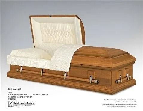 Open wooden casket with a cream-colored interior and bedding, displayed in a well-lit setting.