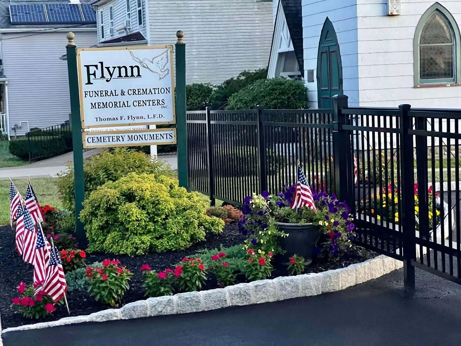 Sign for Flynn Funeral Home with flowers, American flags, and black fence.
