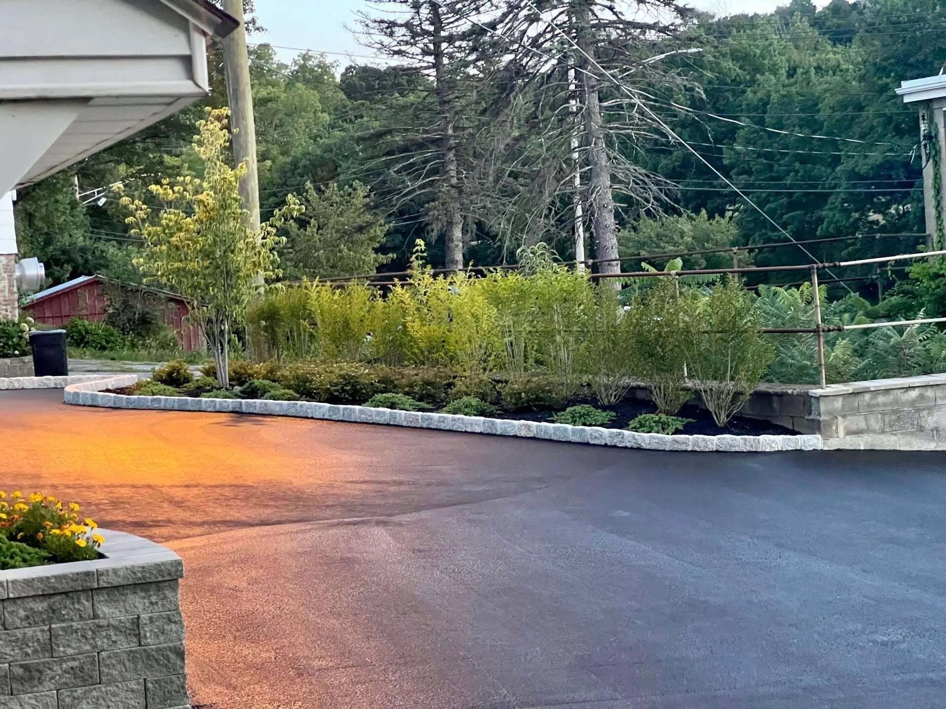 Asphalt driveway with a stone wall and landscaping. Trees and building in the background.