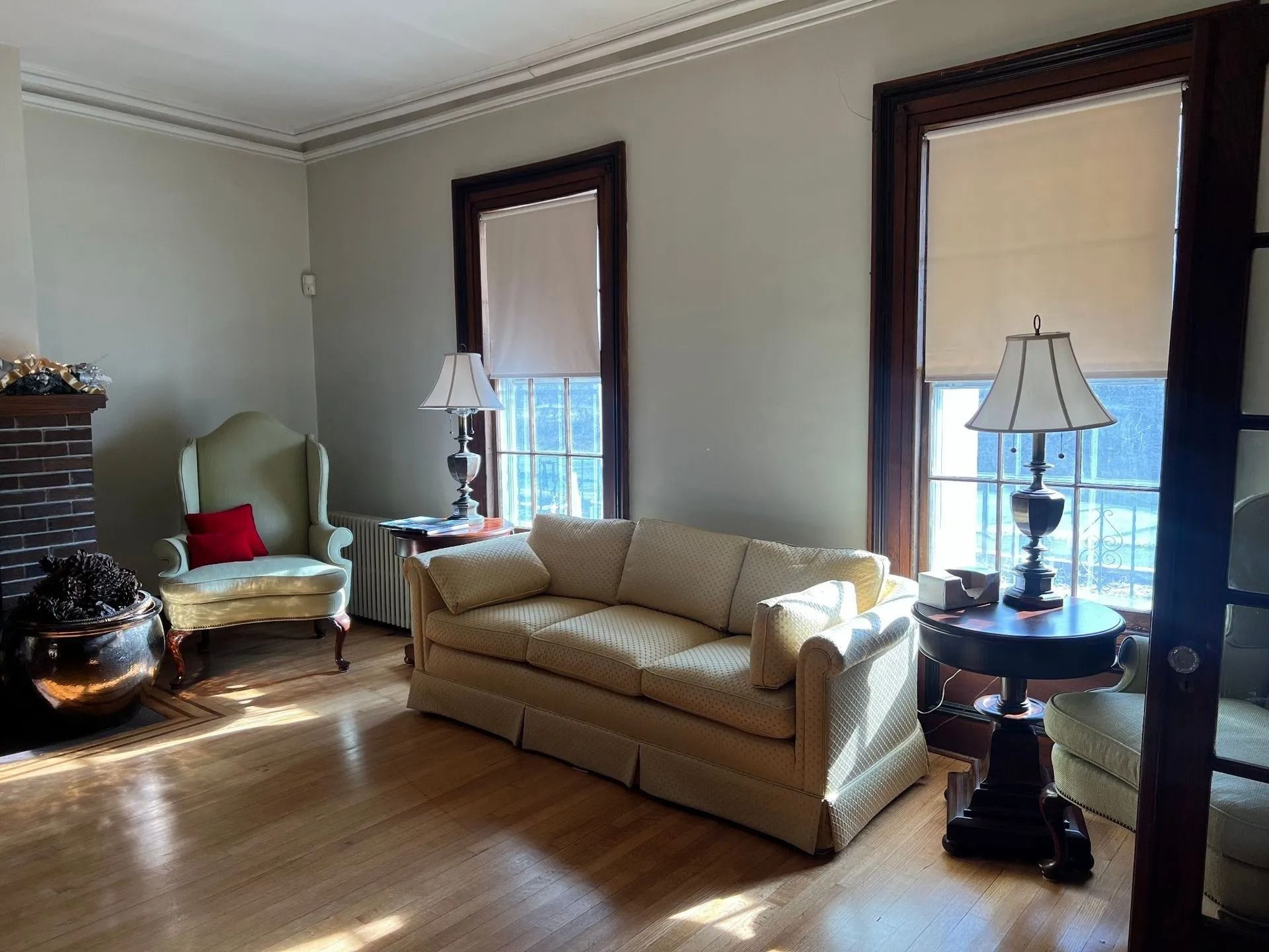Living room with sofa, armchair, two lamps, windows, and fireplace. Sunlight on wood floor.