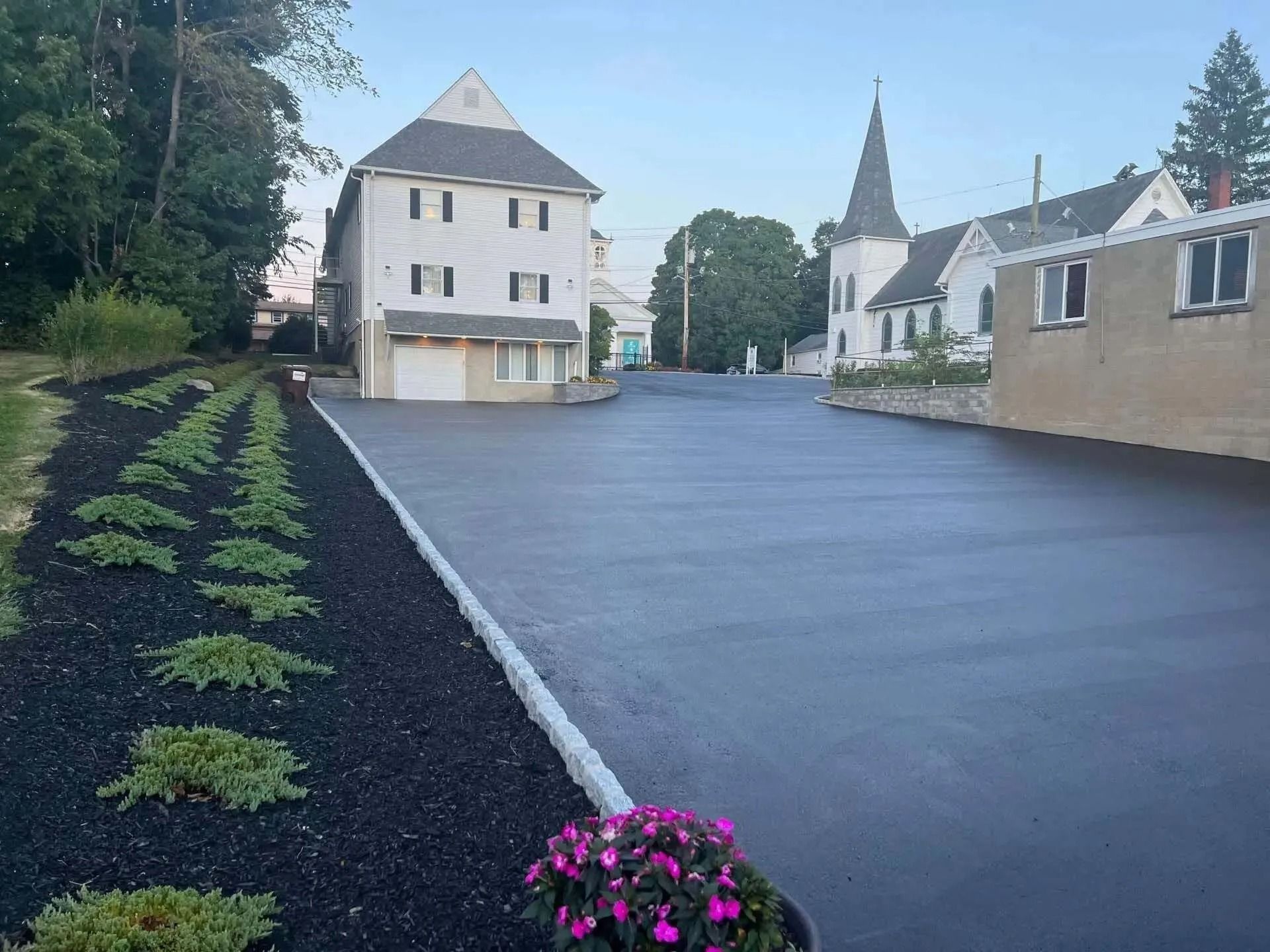 Asphalt lot with a row of green plants against black mulch. Buildings and a church in the background.