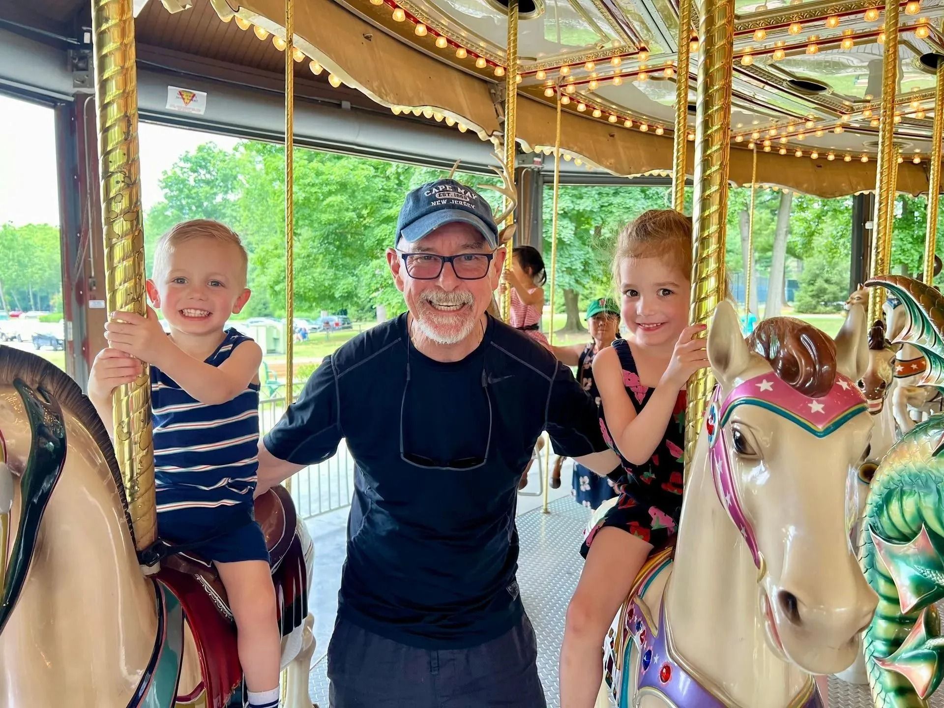 Man smiles between two children on carousel horses; outdoors, green trees visible.