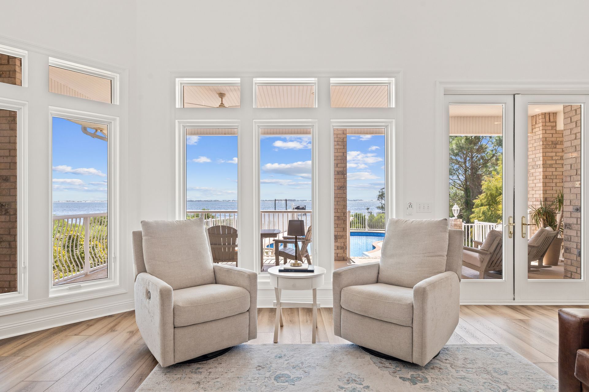 Two beige armchairs face large windows overlooking a deck, pool, and distant blue sky.