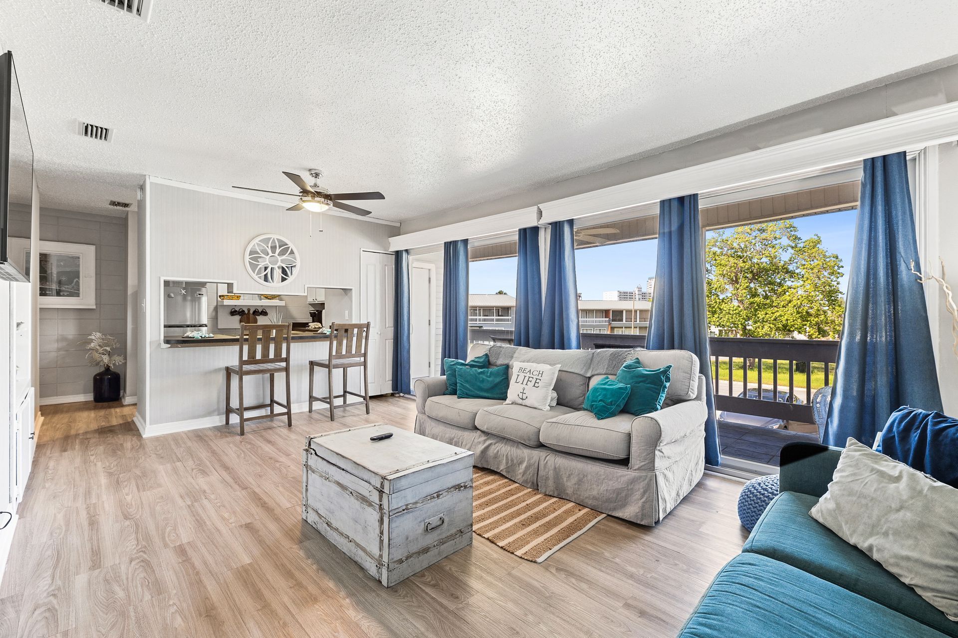 Cozy living room with gray couch, teal pillows, and blue curtains, opening to a balcony.