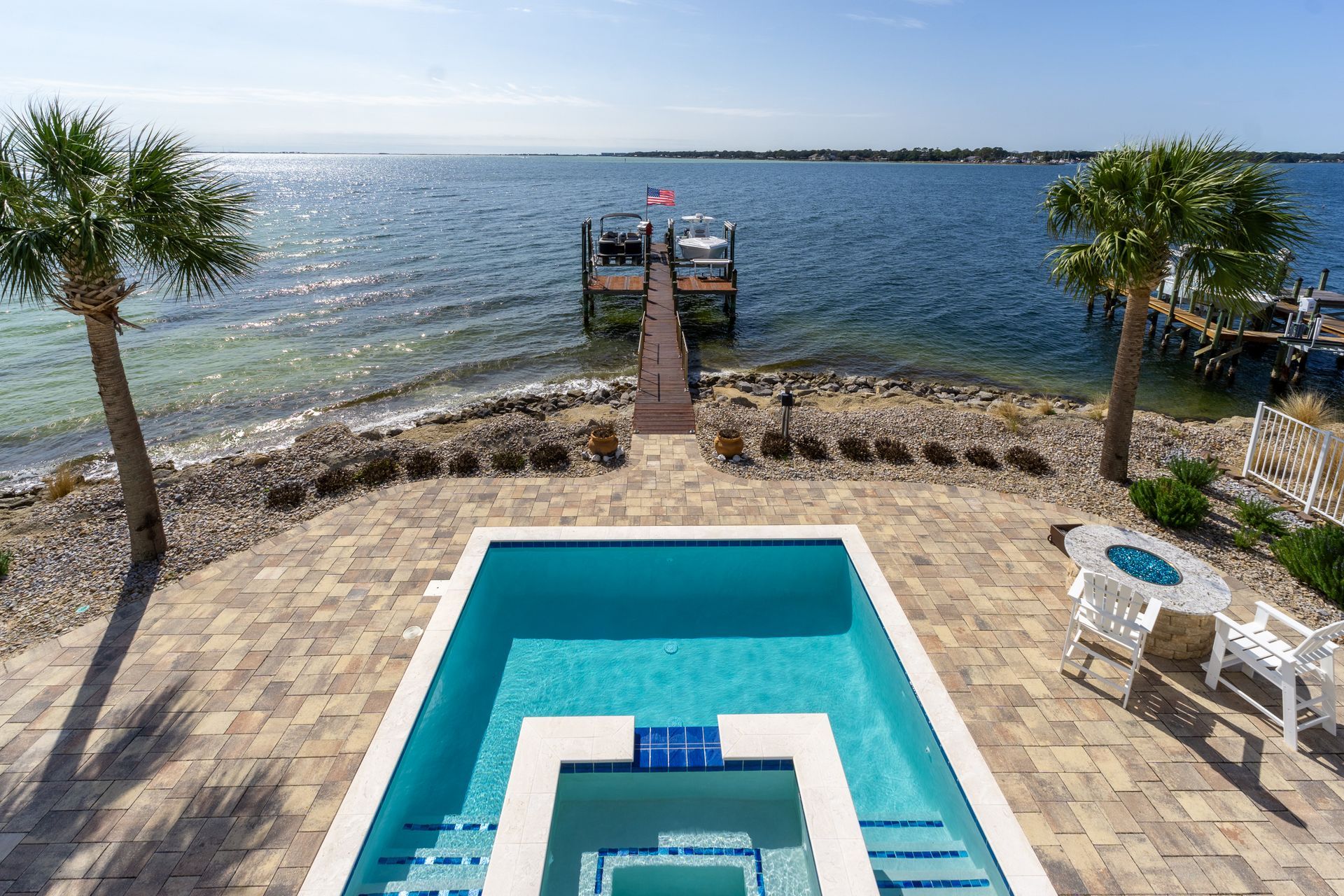 Pool with steps leading to the water and a dock with boats in the distance under a sunny sky.