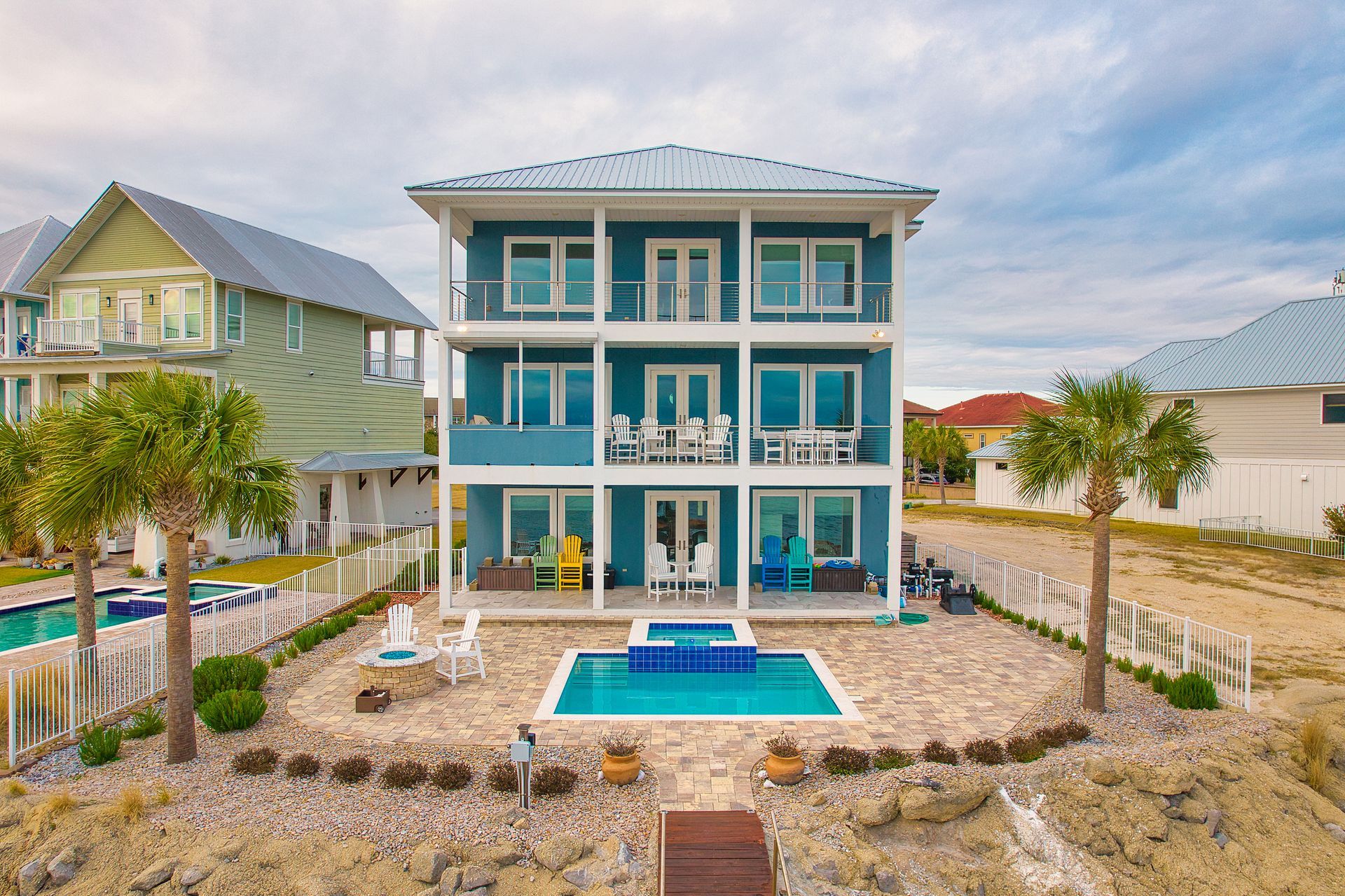 Blue three-story beach house with a pool, palm trees, and white trim, next to other houses on a beach.