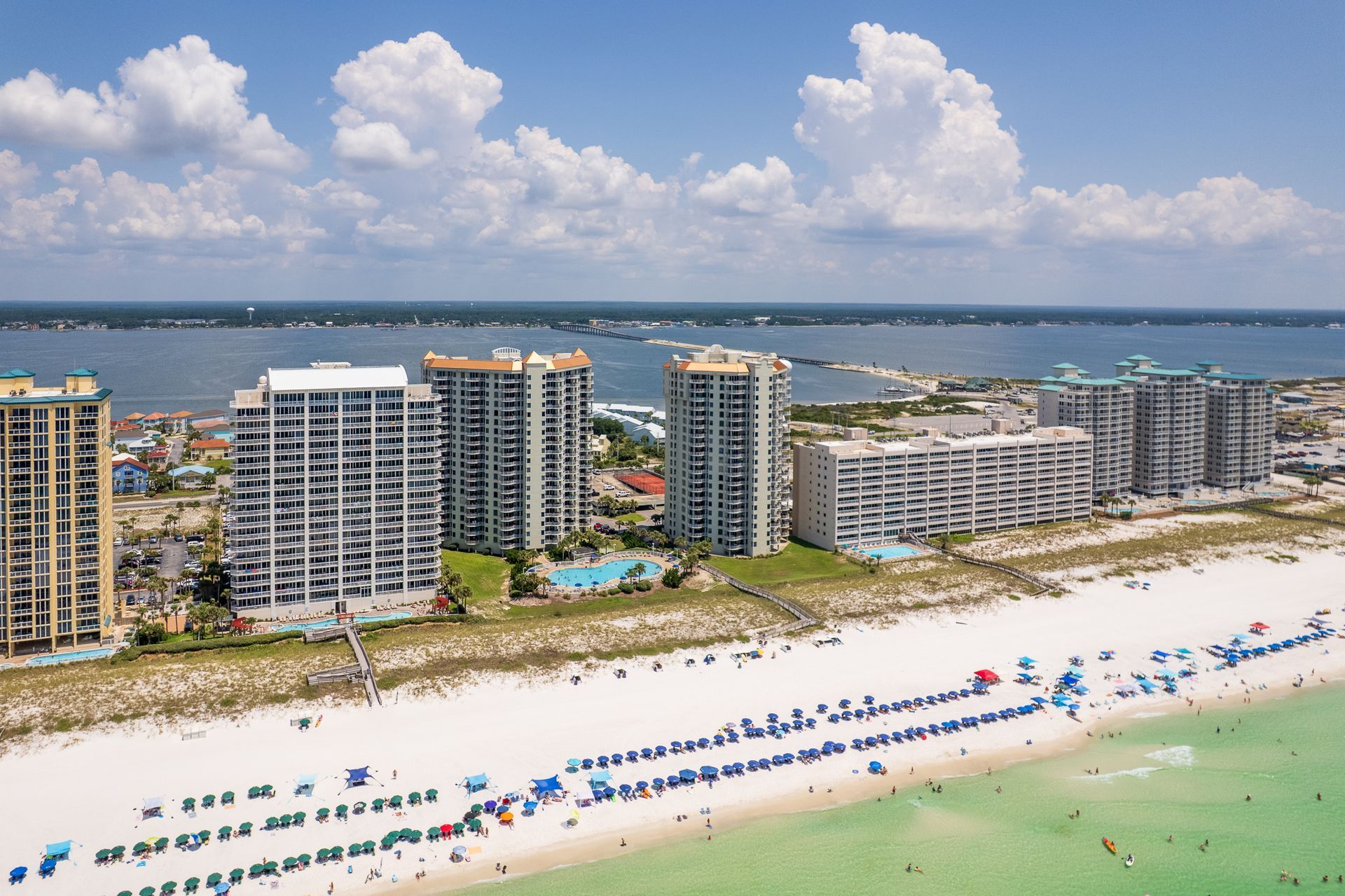 Beach with white sand and turquoise water; high-rise condos line the shore under a blue sky with puffy clouds.