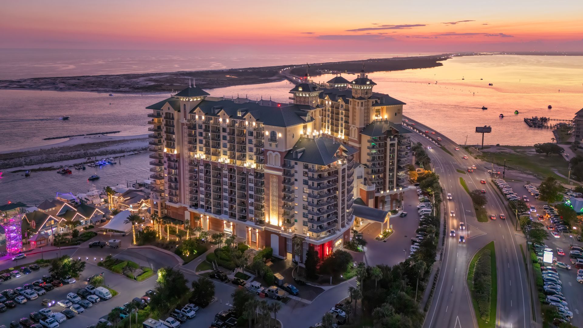 Luxury resort at sunset overlooking beach and waterway with cars on nearby highway.