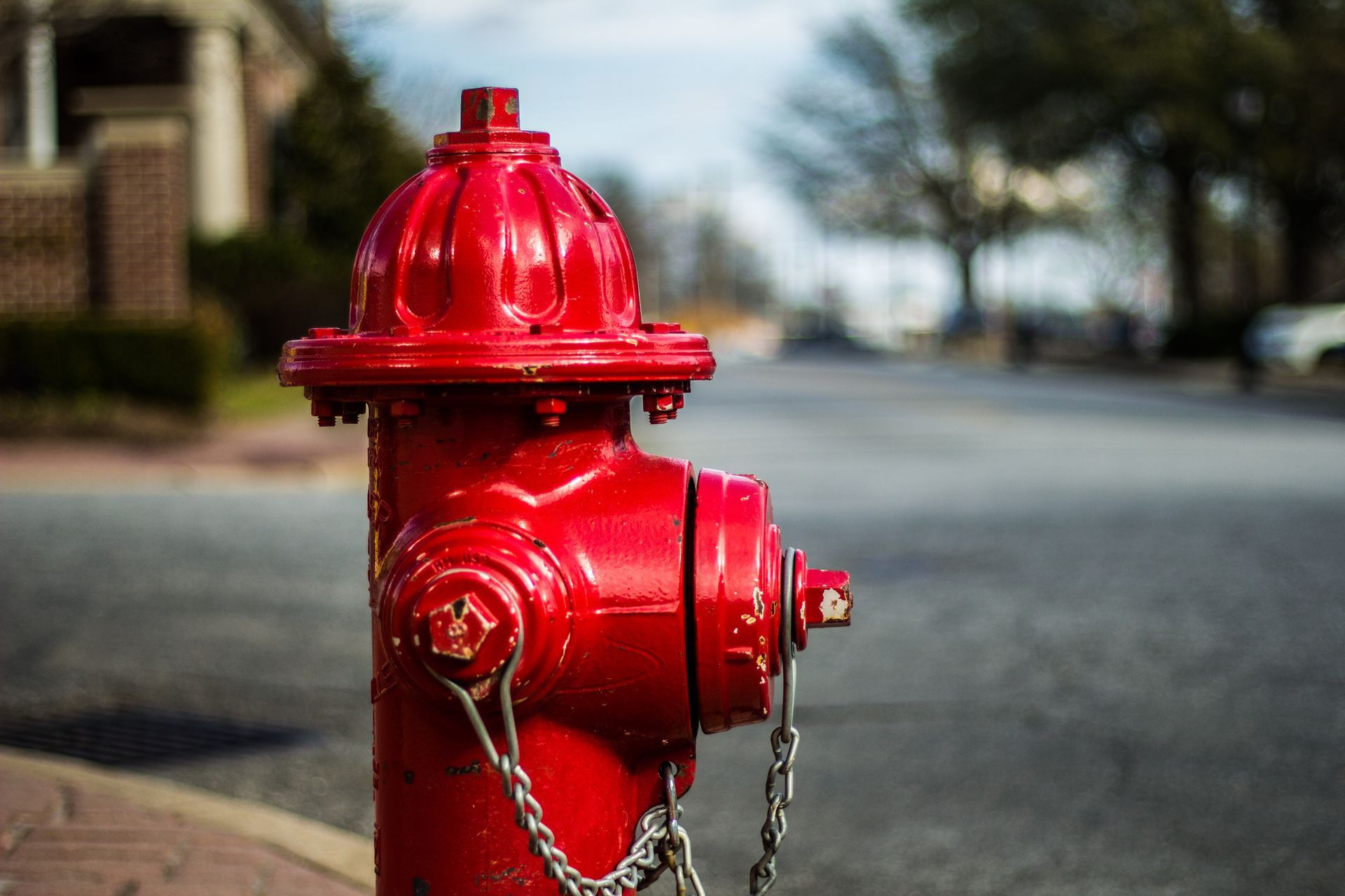 Bright red fire hydrant standing on a street corner with buildings and trees in the background.