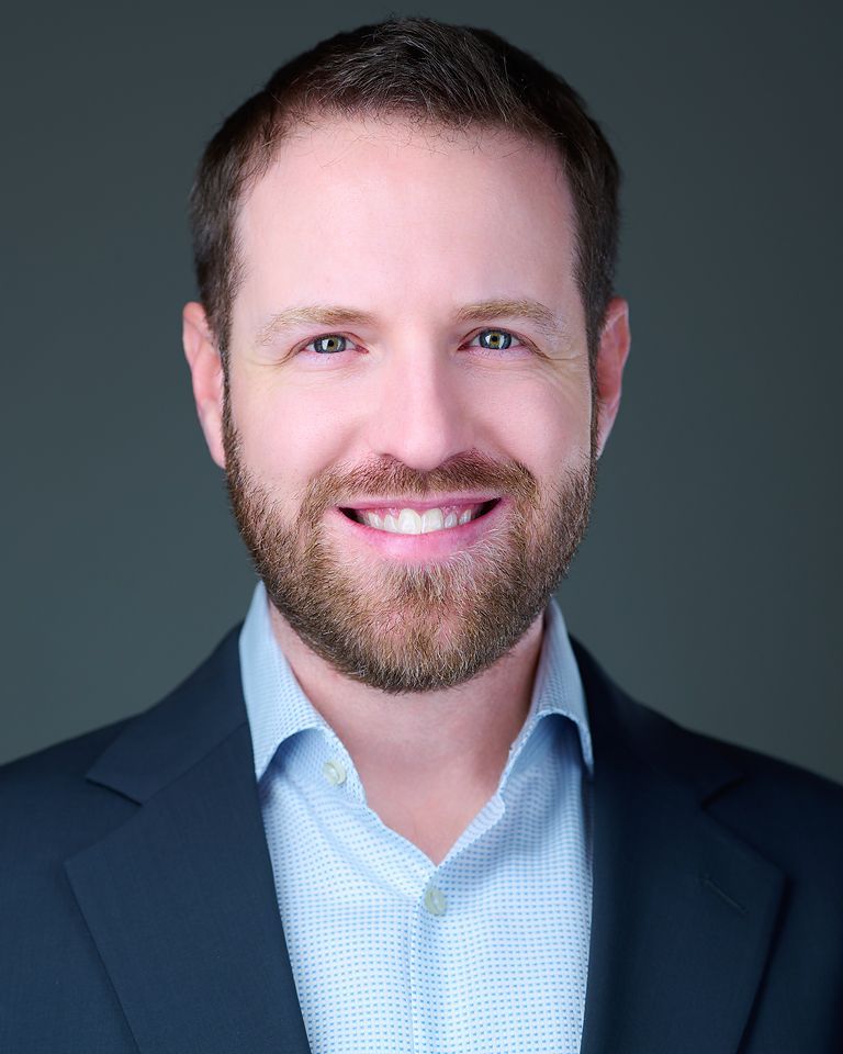 A man with a beard is wearing a white shirt and smiling.