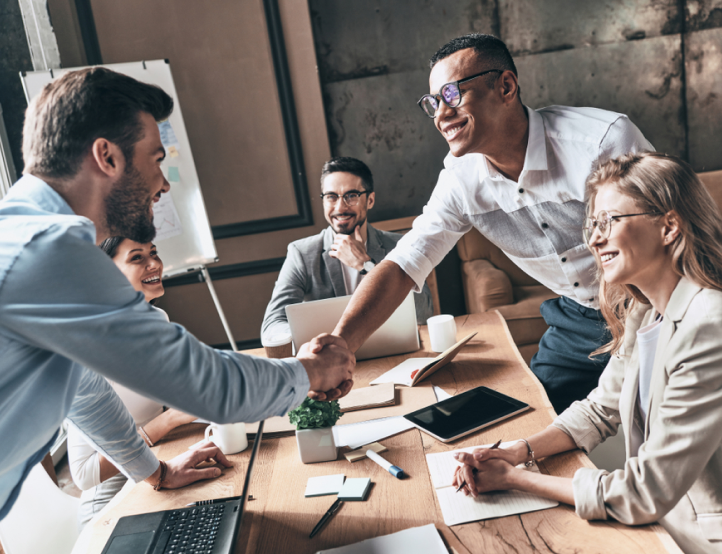 A man is shaking hands with a woman while a group of people are sitting around a table.