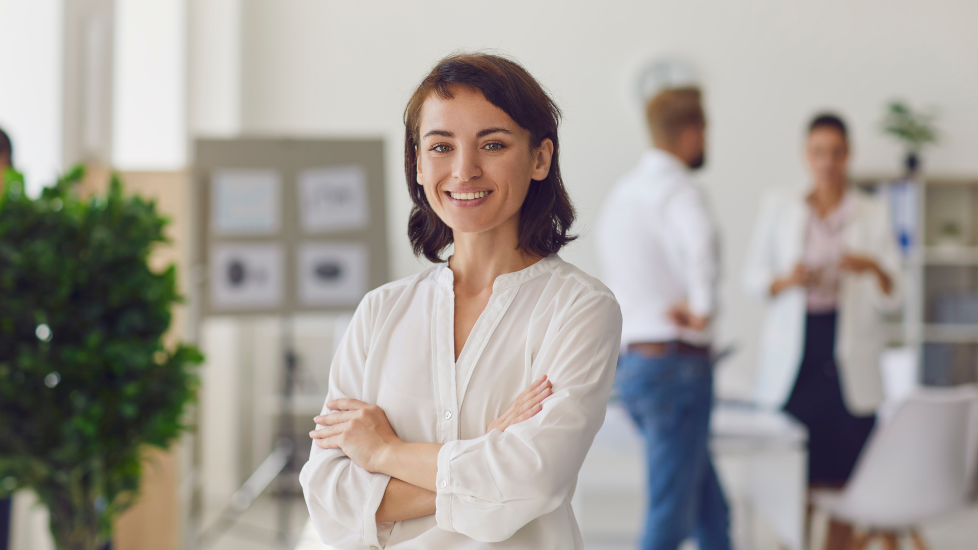 A woman is standing in an office with her arms crossed and smiling.
