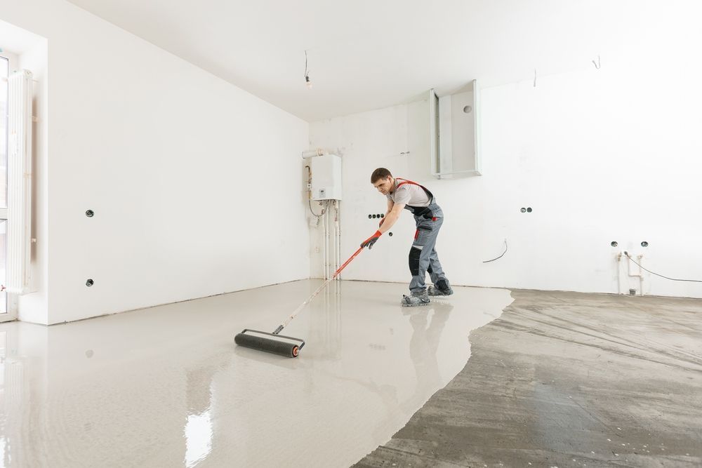 Person applying a light-colored epoxy floor coating with a roller in a room with white walls.