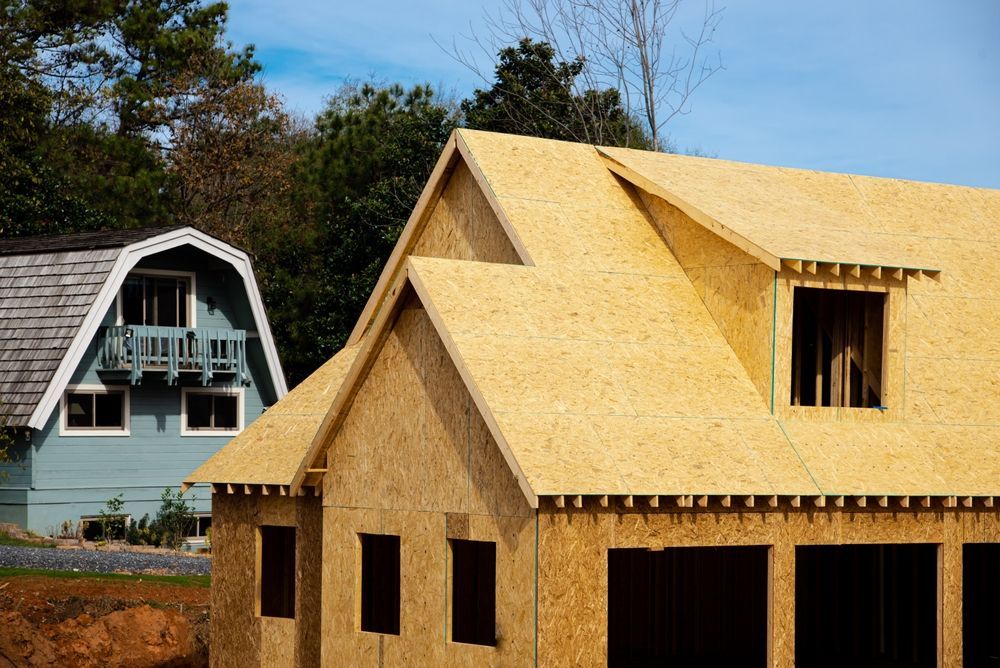 House under construction next to a finished blue house, with plywood roof and clear blue sky.