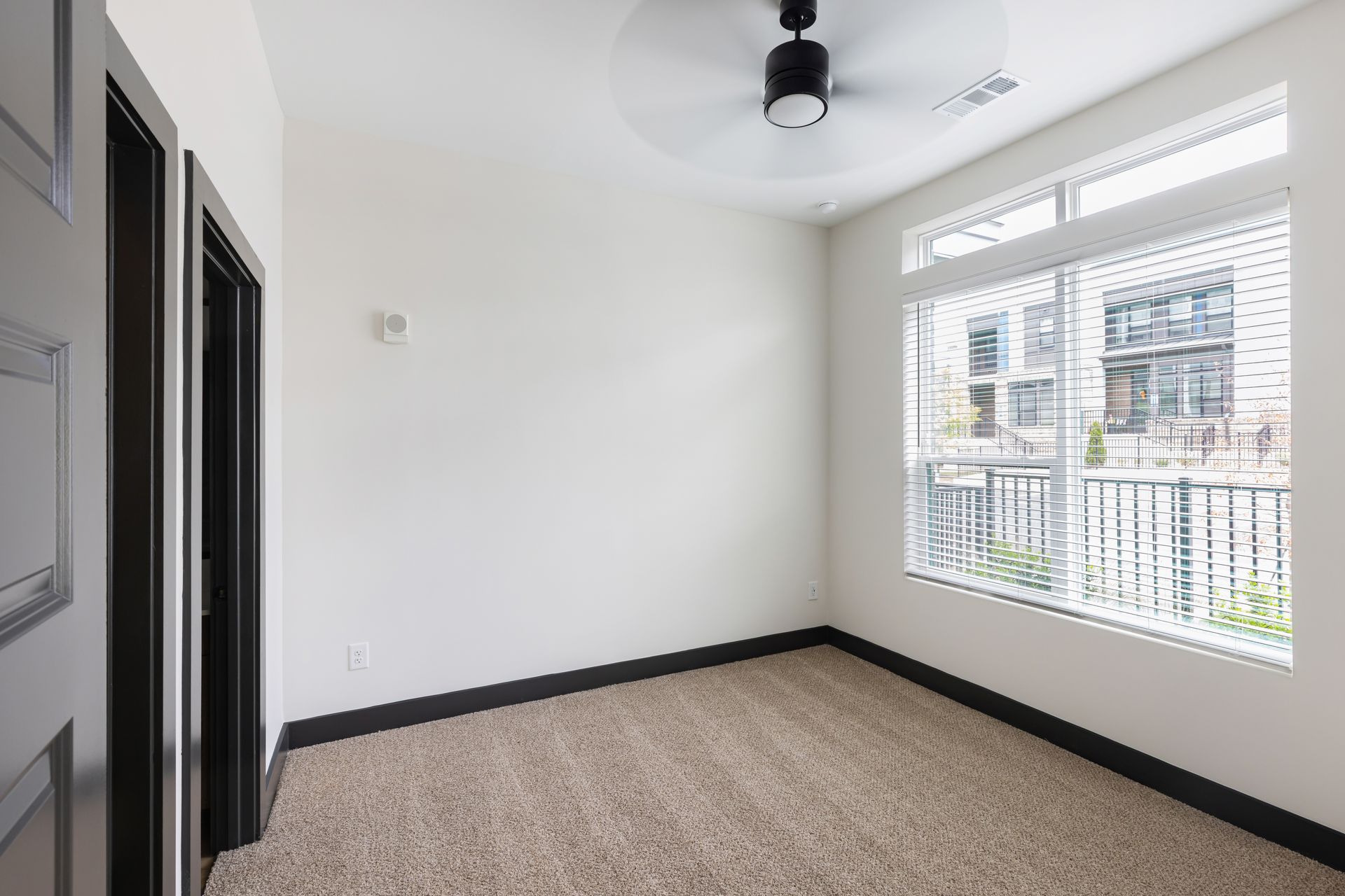 Empty carpeted room with white walls, dark trim, ceiling fan, and large window overlooking balconies at Vela Park apartments in East Lake, Atlanta, GA.